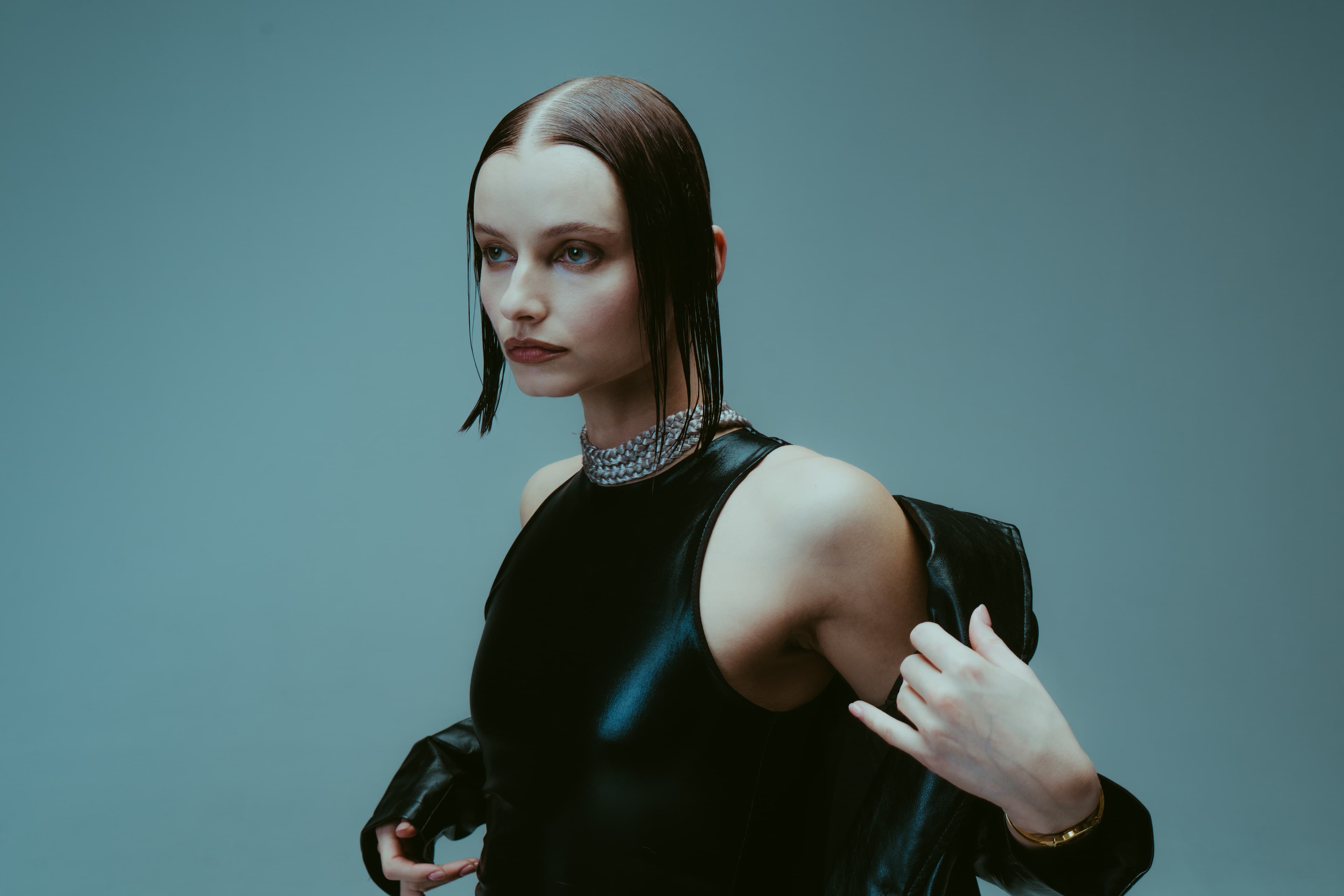 A woman with slicked-back hair and a crystal choker in a black leather top against a teal backdrop