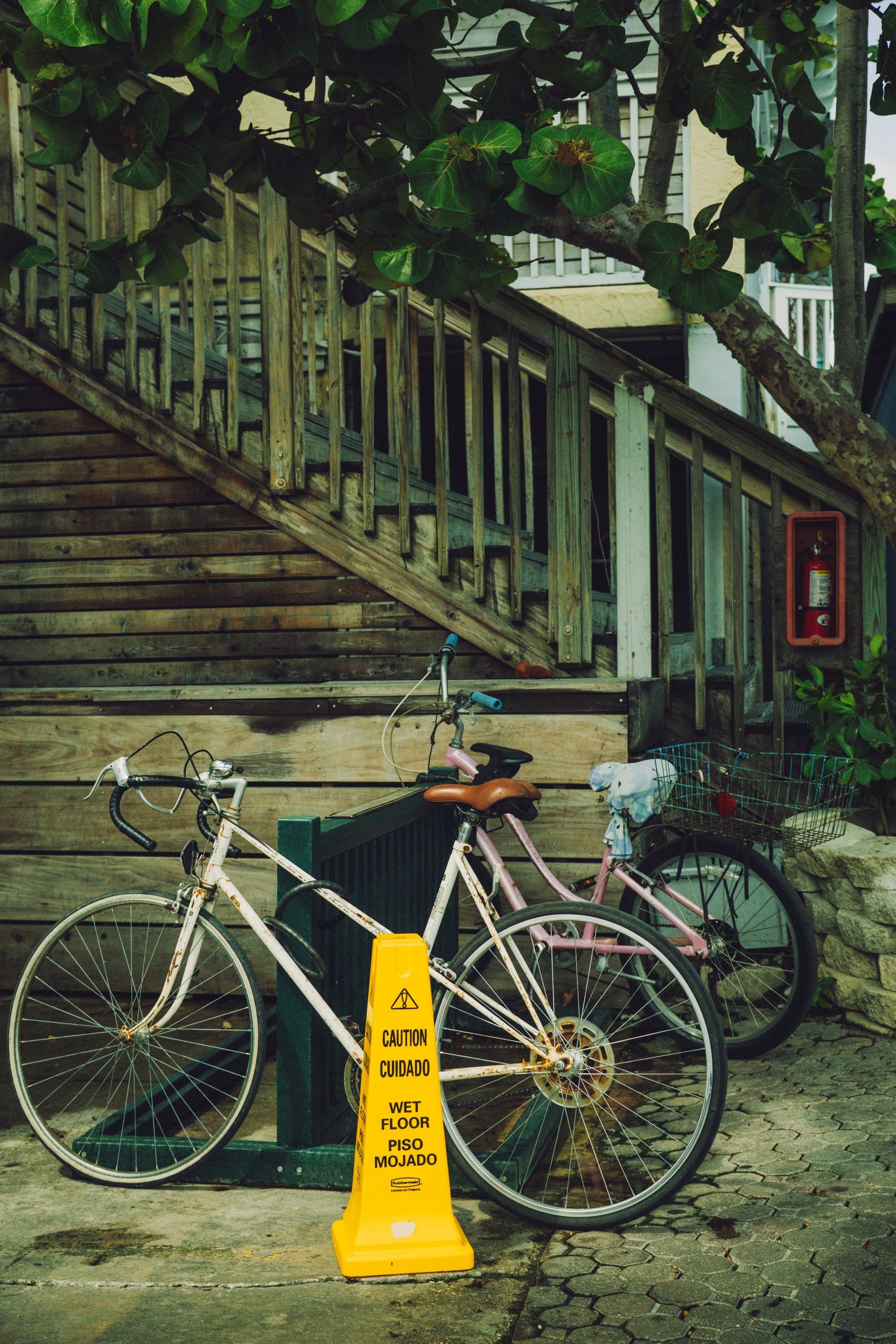 Two bicycles parked under weathered wooden stairs beside a yellow wet-floor sign