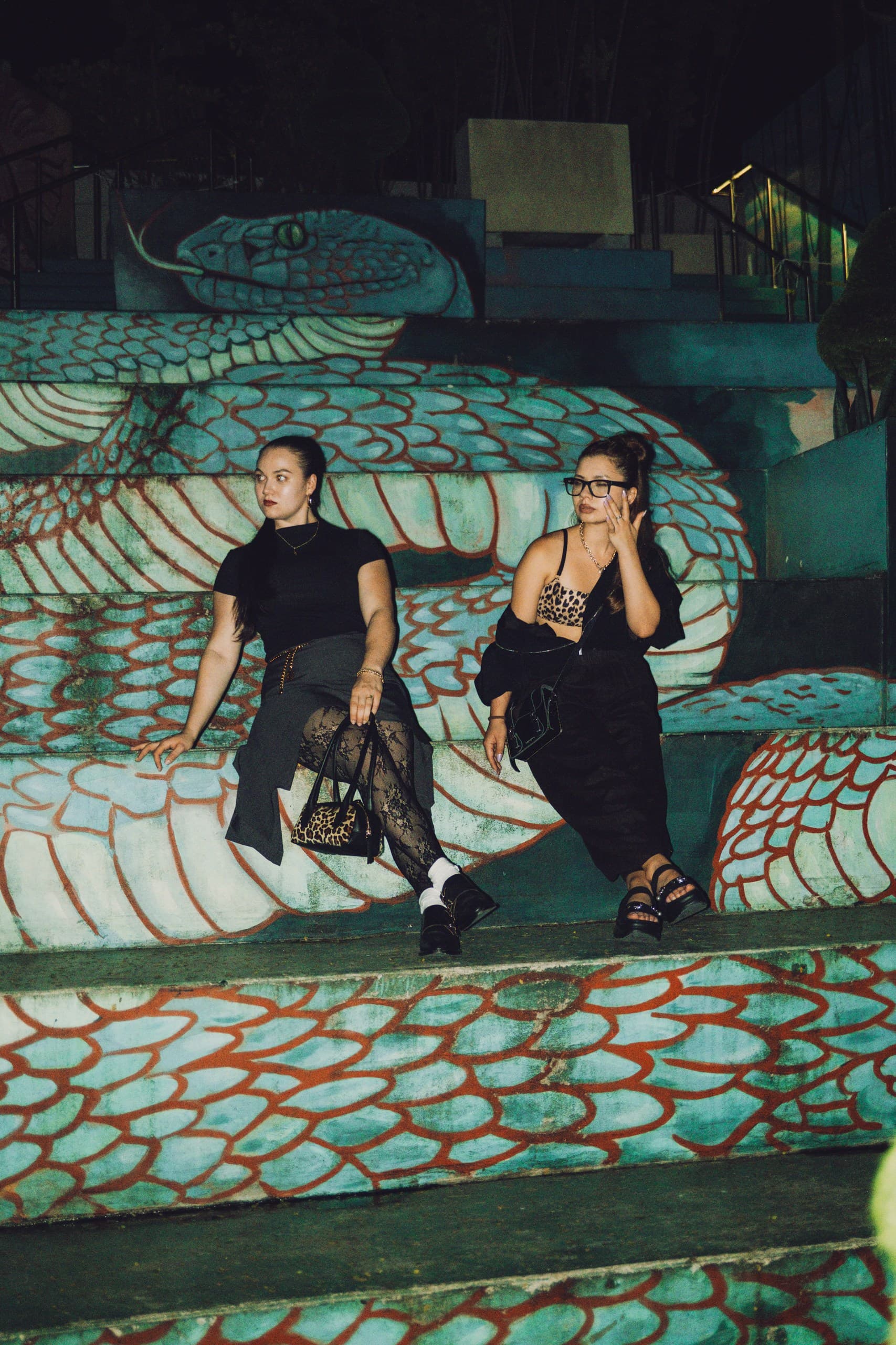 Two women in black sitting on steps painted with a turquoise and red serpent mural