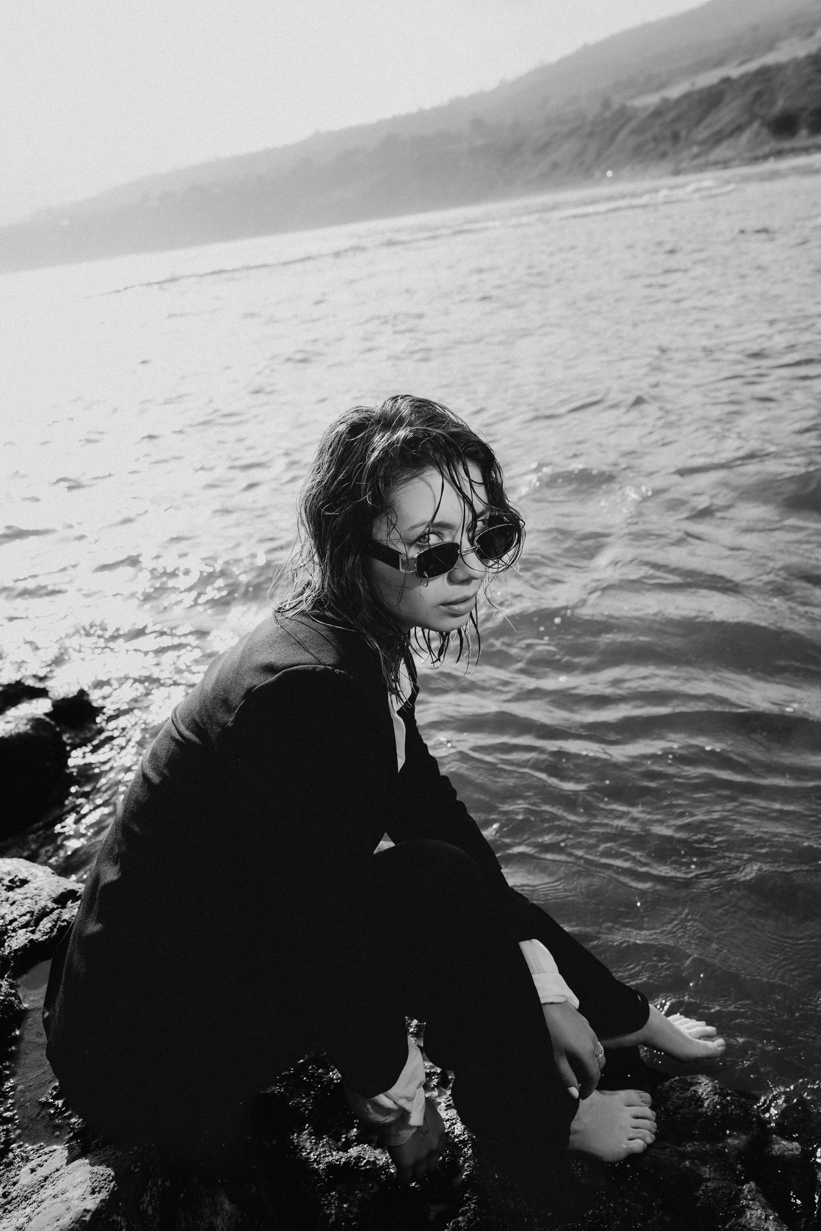 Black-and-white photo of a woman in sunglasses and a dark coat crouching in the surf