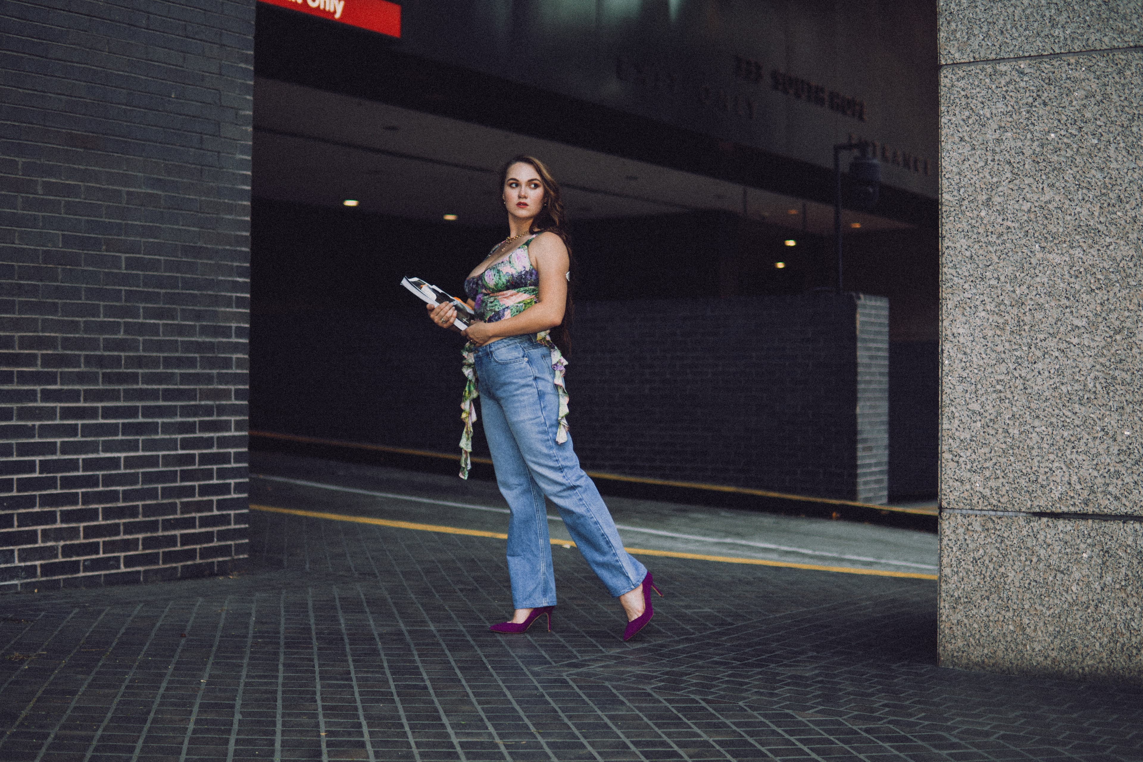 A woman in a floral corset top and baggy light-wash jeans walking past a parking garage at night, a magazine in her hand