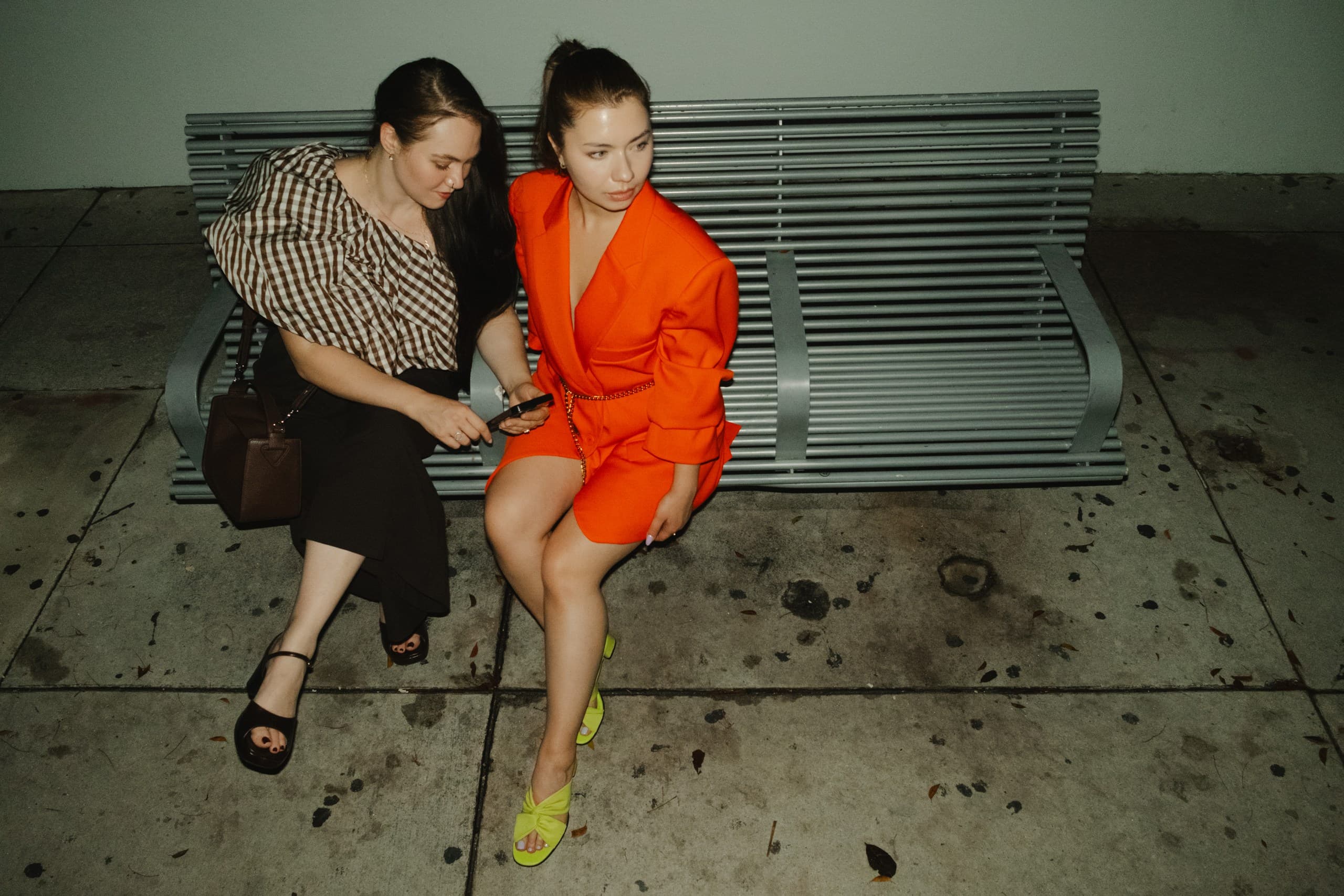Two friends on a metal bench at night looking at a phone, one in a striped top, one in a bright orange suit and lime heels