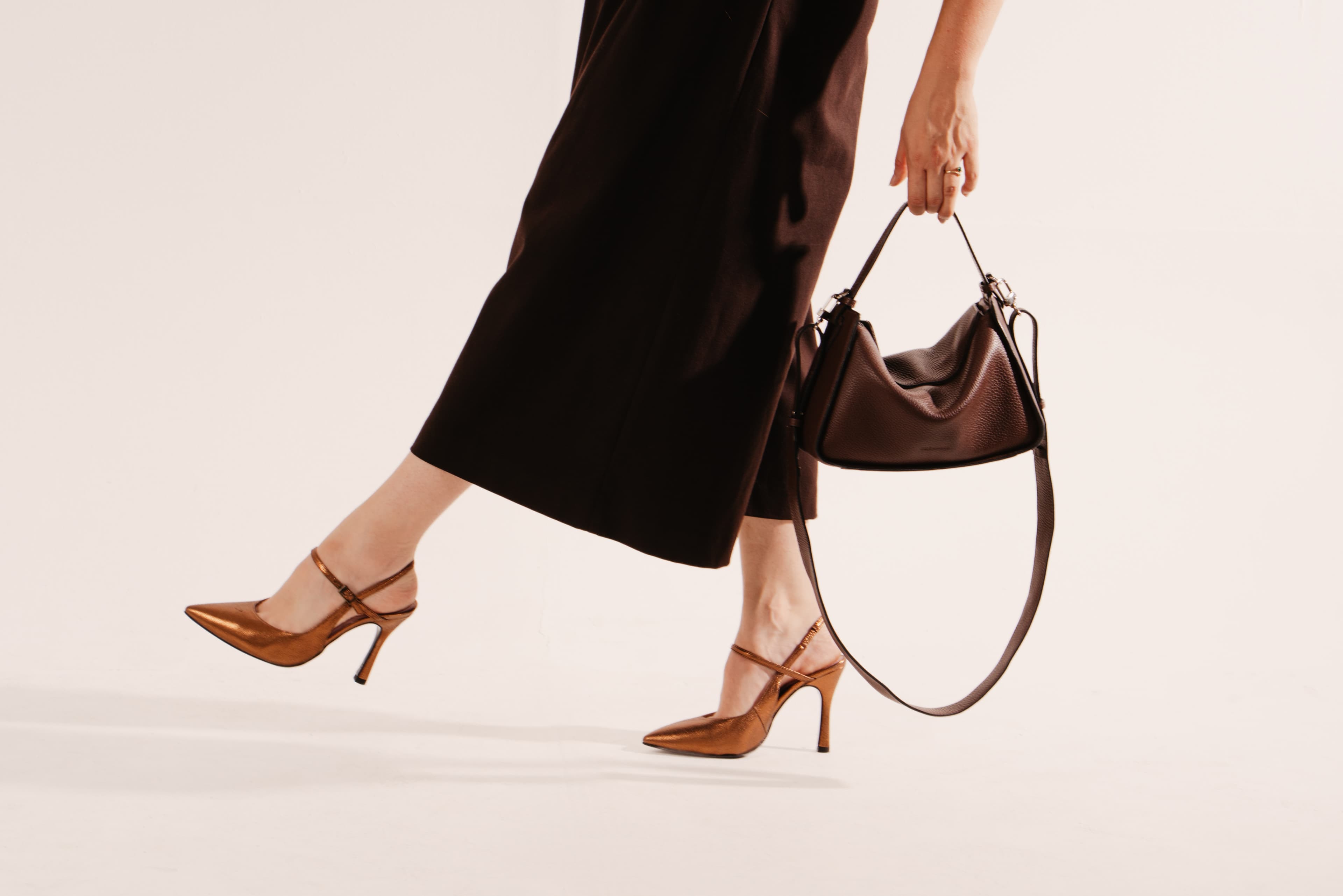 Mid-stride legs in black cropped trousers and tan slingback heels, a dark brown leather shoulder bag swinging from one hand