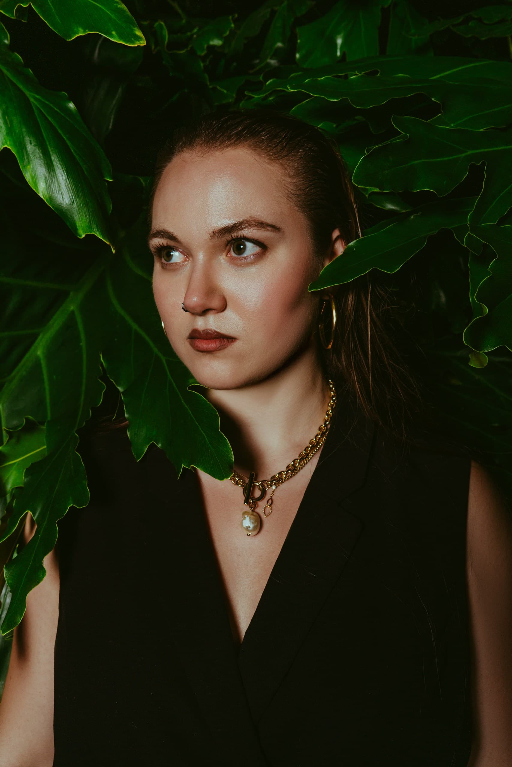 A woman with slicked-back hair and gold chains framed by large green leaves