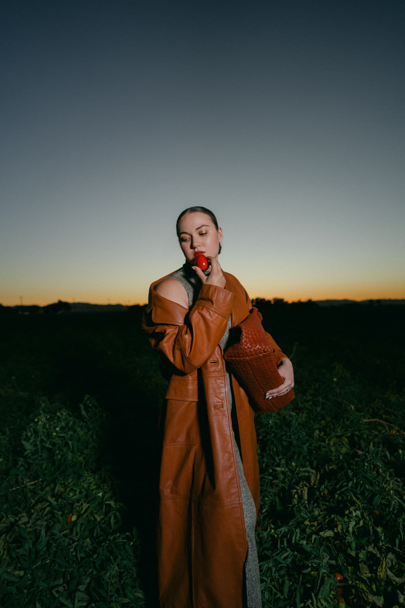 A woman in a cognac leather coat holding a tomato to her lips in a field at sunset