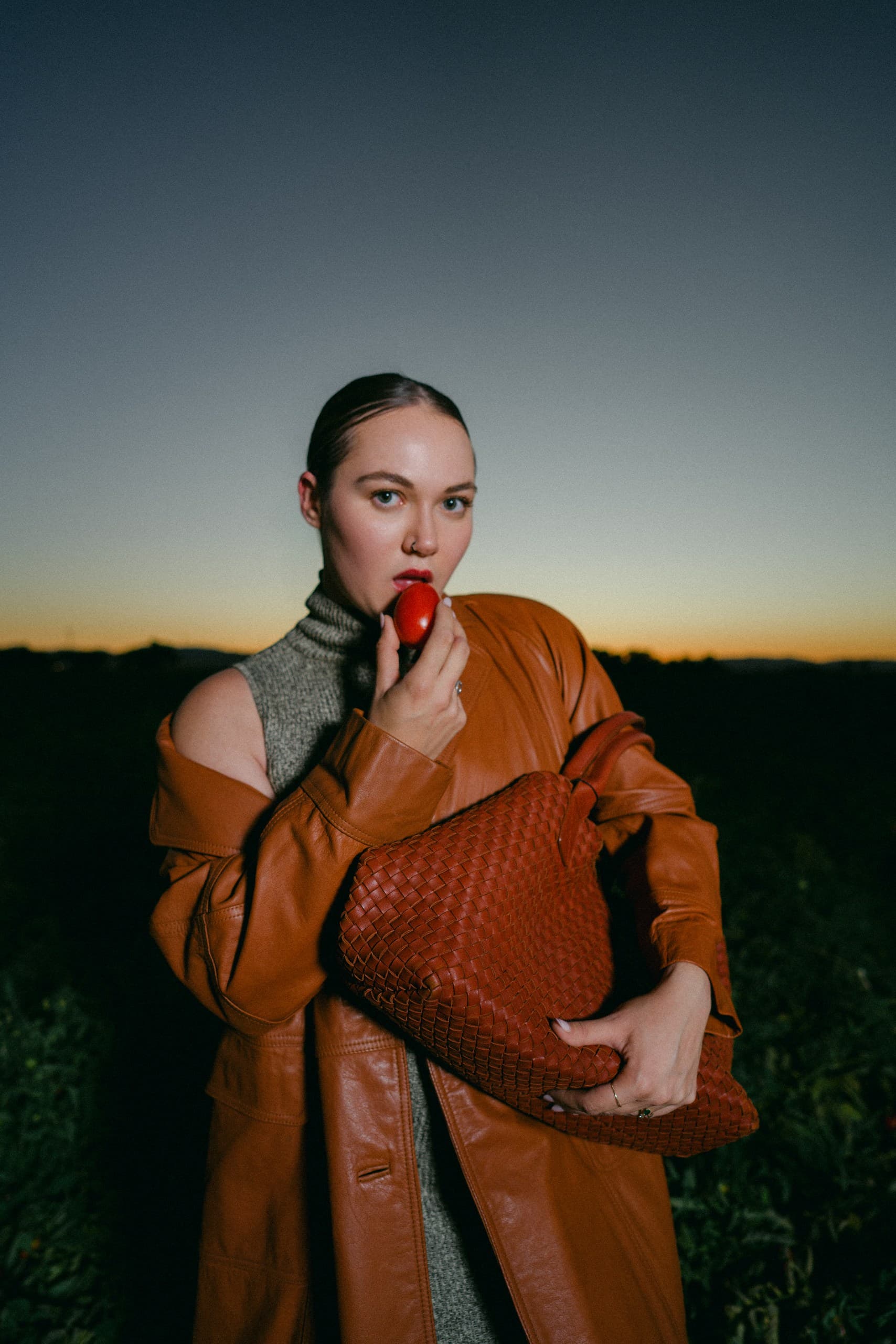 A woman in a cognac leather coat and grey turtleneck biting a tomato at dusk, a woven tote under her arm
