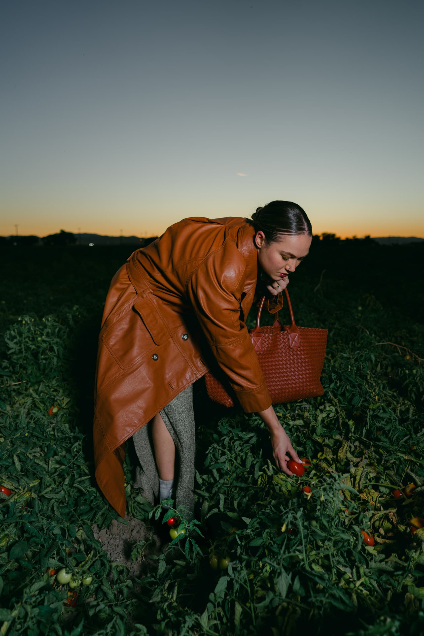 A woman in a long cognac leather coat bending to pick tomatoes in a field at dusk, a woven tote on her shoulder