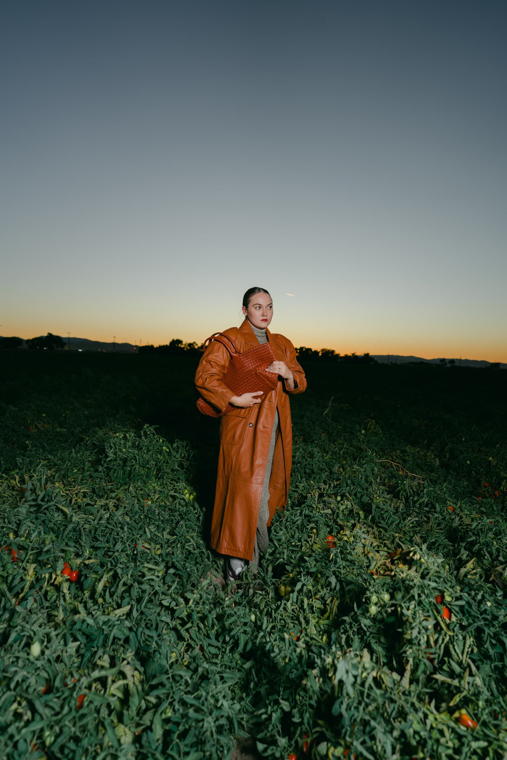 A woman in a long cognac leather coat standing in a tomato field under a dusk sky