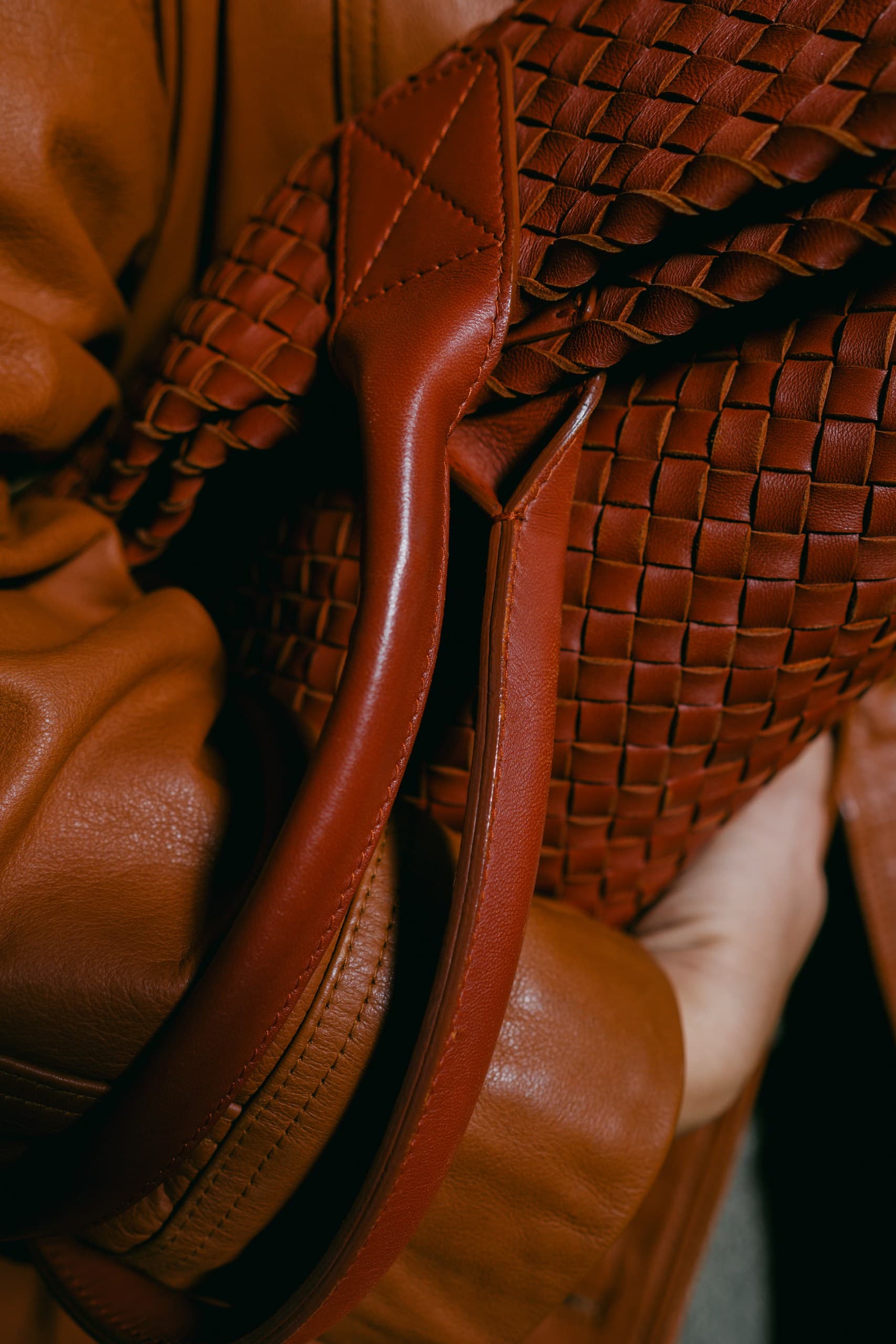 Close-up of the rolled leather handle of a cognac woven tote against a leather coat