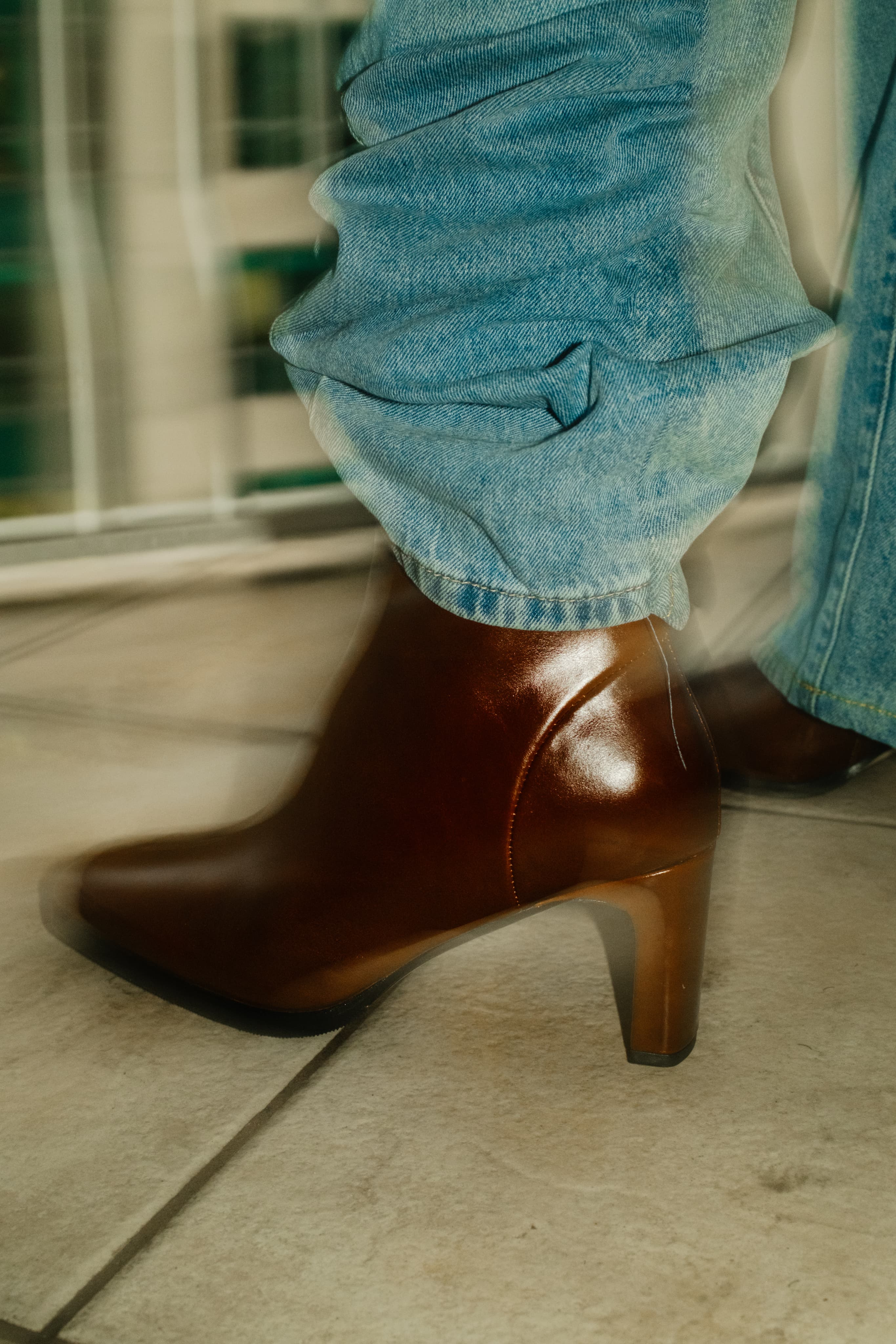 A brown leather heeled ankle boot mid-step beneath light-wash jeans