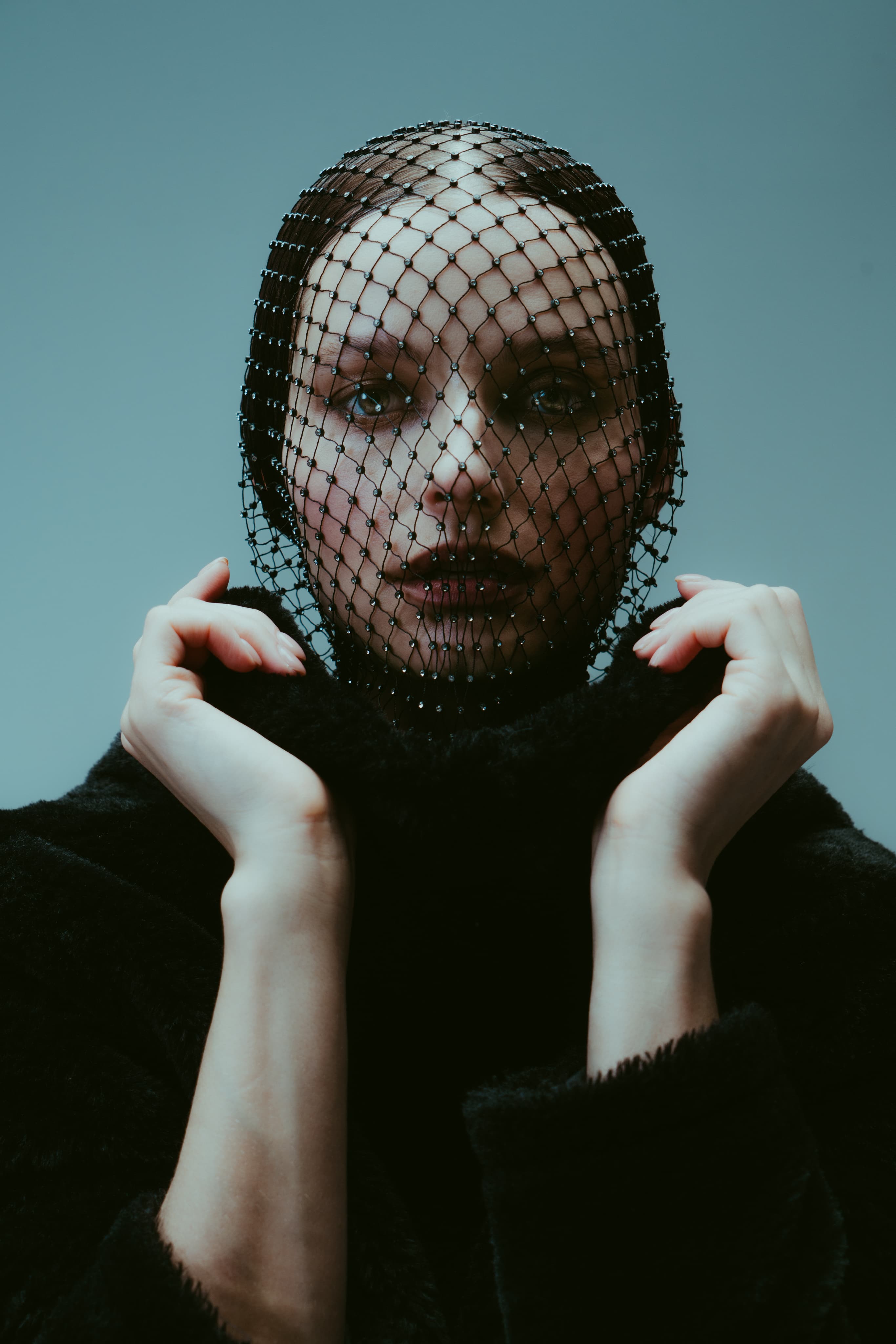 A woman in a black fishnet hood and black coat holding up her collar against a blue-grey backdrop