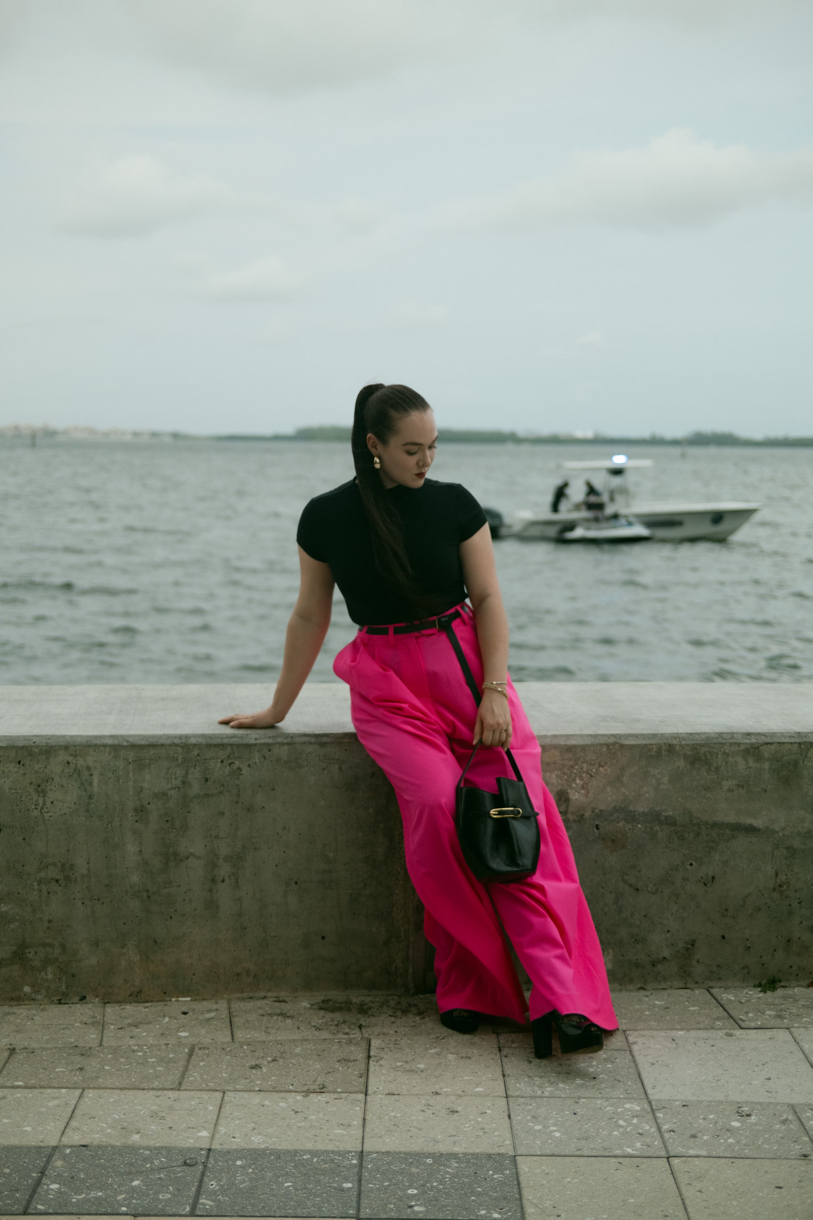 A woman in a black top and hot-pink wide trousers leaning on a seawall, a boat on the water behind her