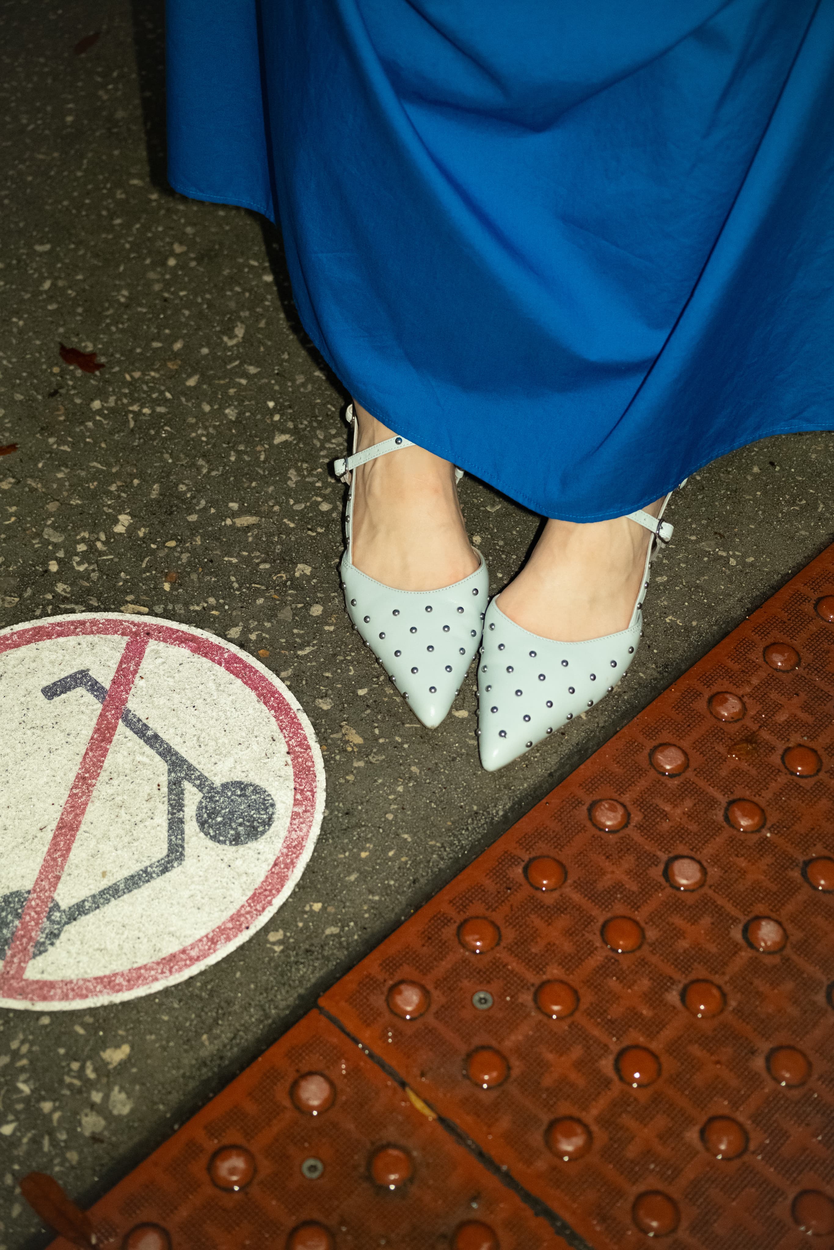 Pale blue studded pointed flats under a cobalt skirt hem on red tiled pavement