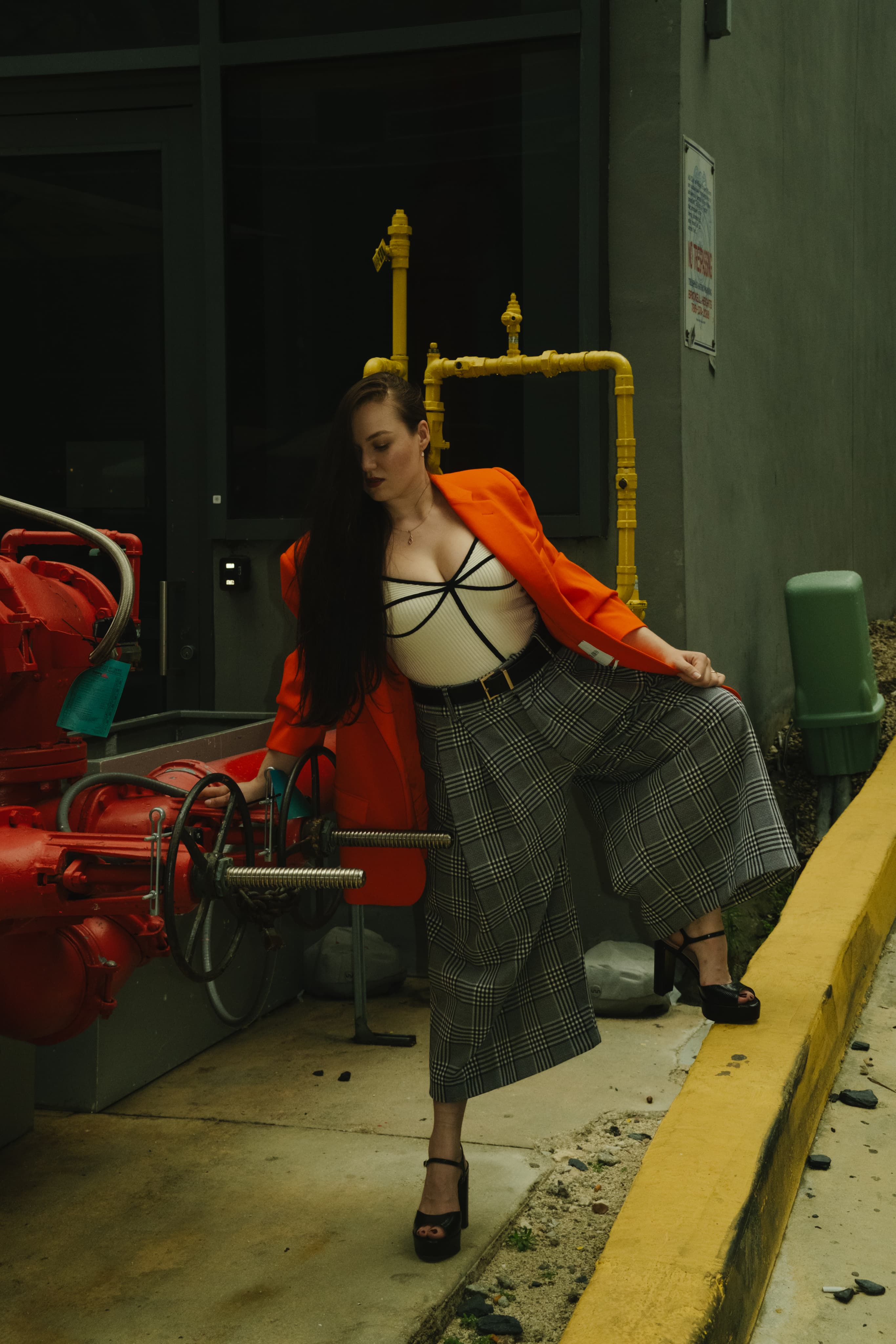 A woman in an orange jacket, black bustier and plaid wide trousers by red industrial pipes