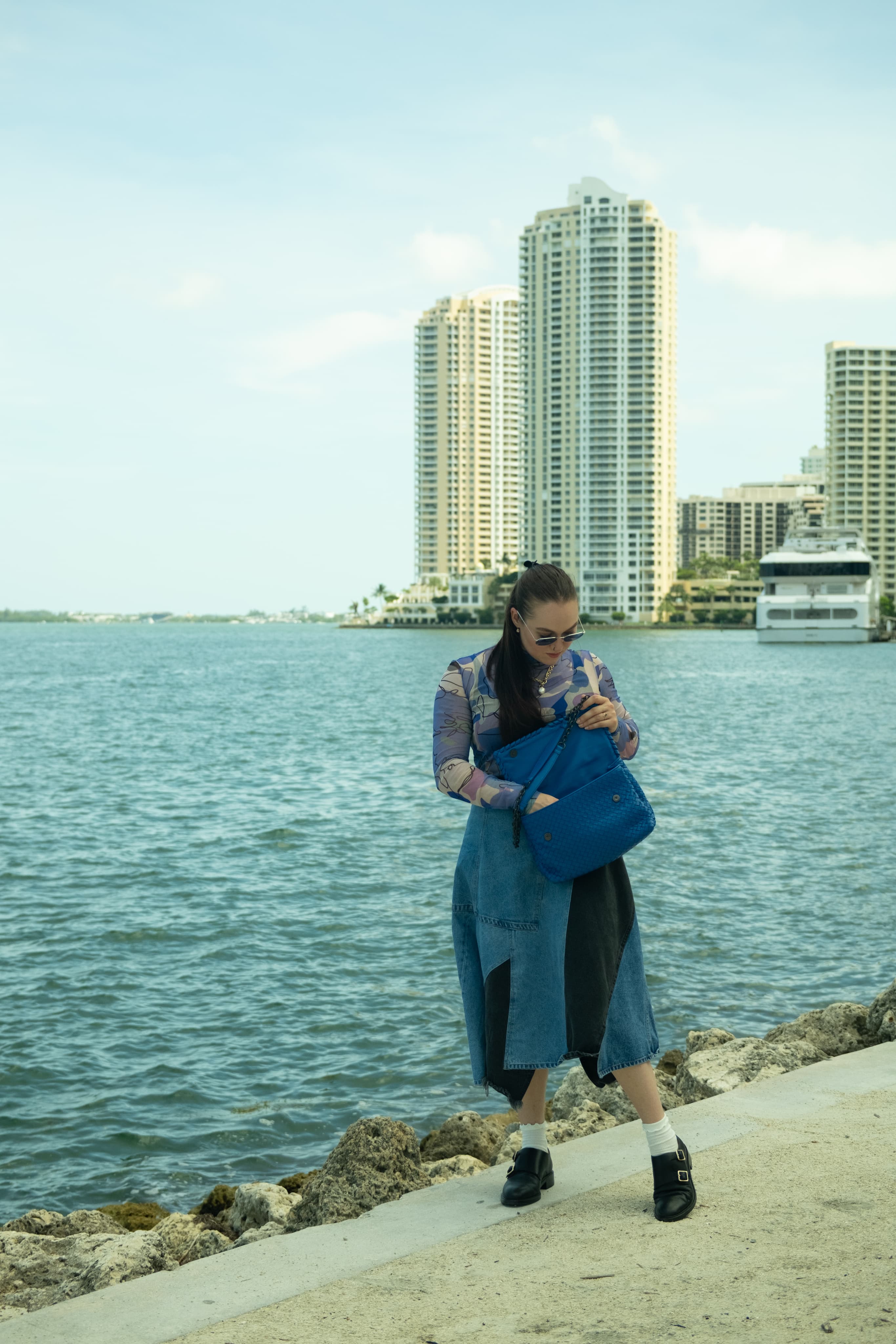 A woman in a long denim coat on a rocky waterfront, high-rise towers across the bay