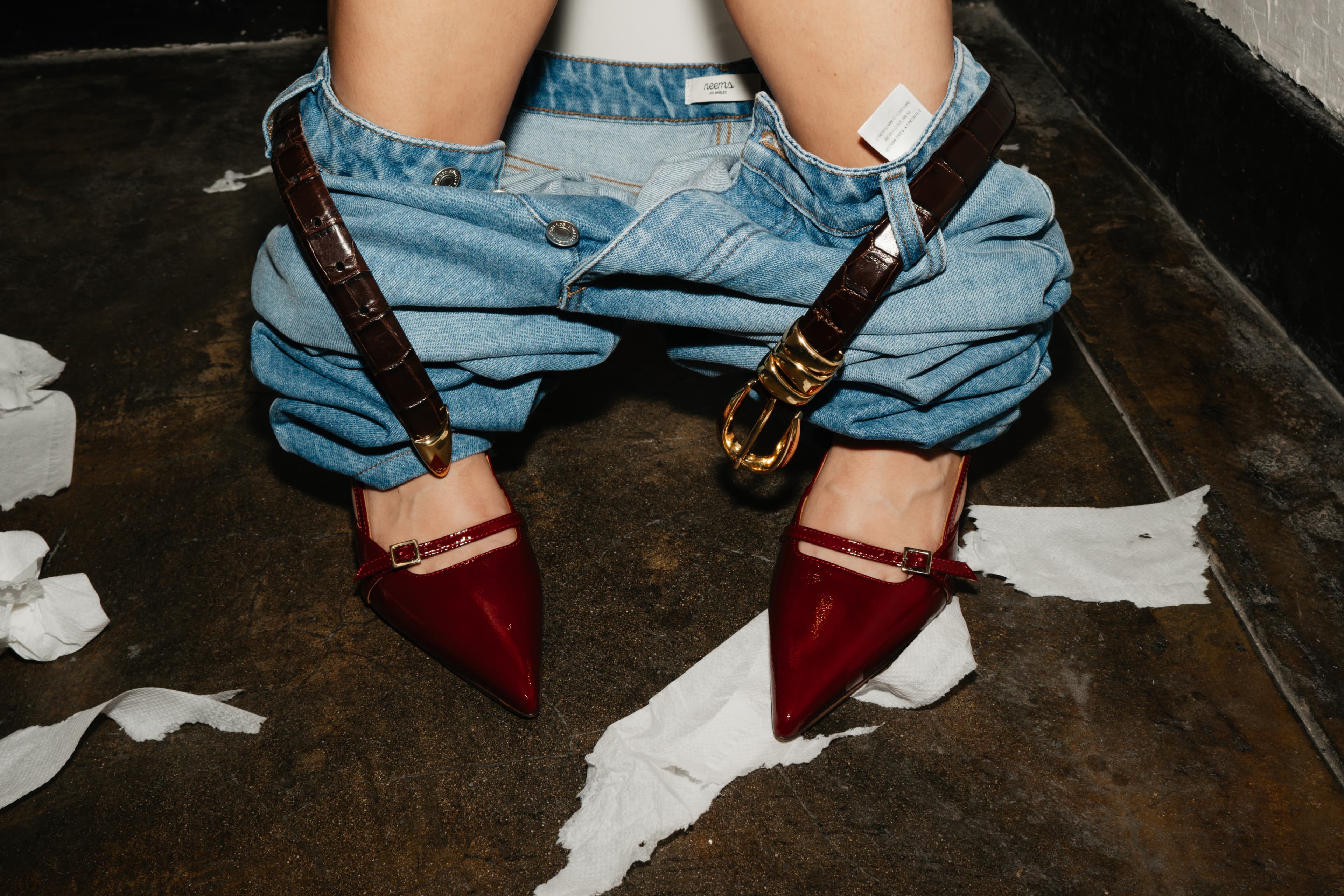 Red patent Mary Jane pumps beneath rolled light-wash jeans and a croc belt with a gold buckle, on a scuffed floor scattered with torn paper