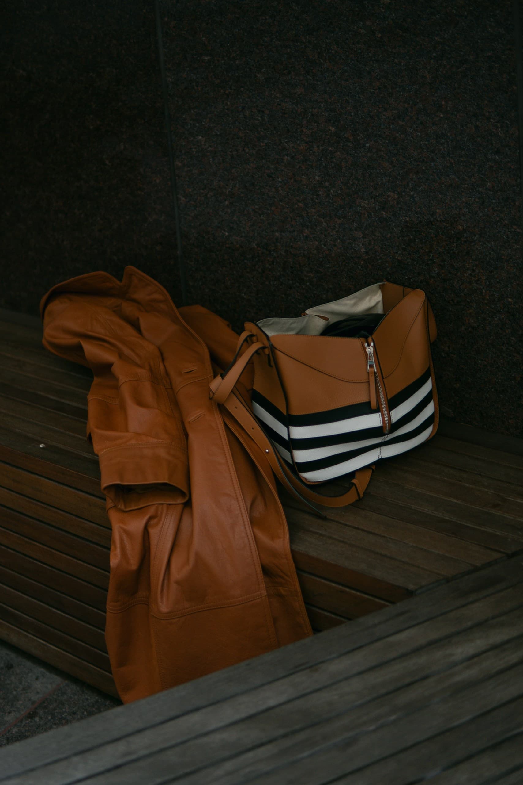 A cognac leather coat lying crumpled beside a striped canvas tote on a wooden deck