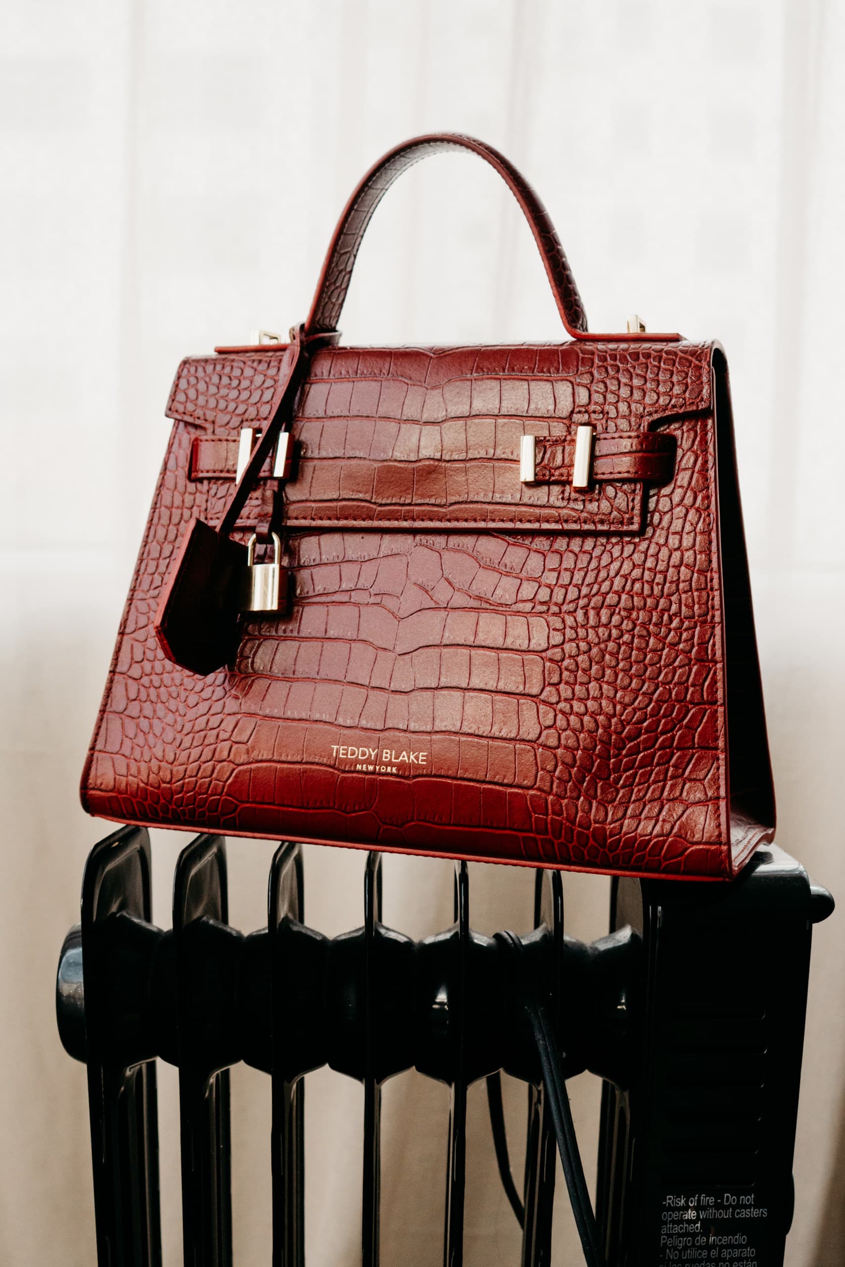 A burgundy croc-embossed top-handle bag with a padlock, set on a black radiator
