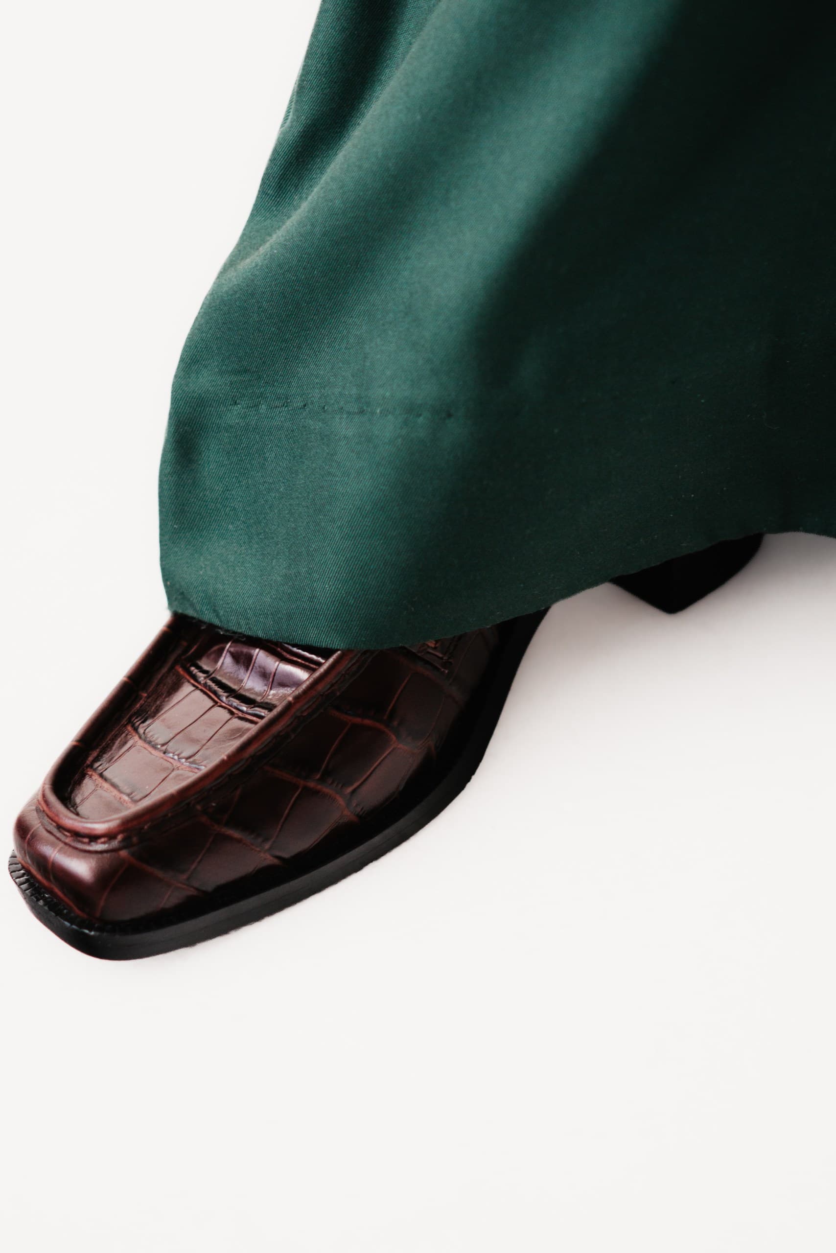 A burgundy croc-embossed square-toe loafer beneath a green trouser hem