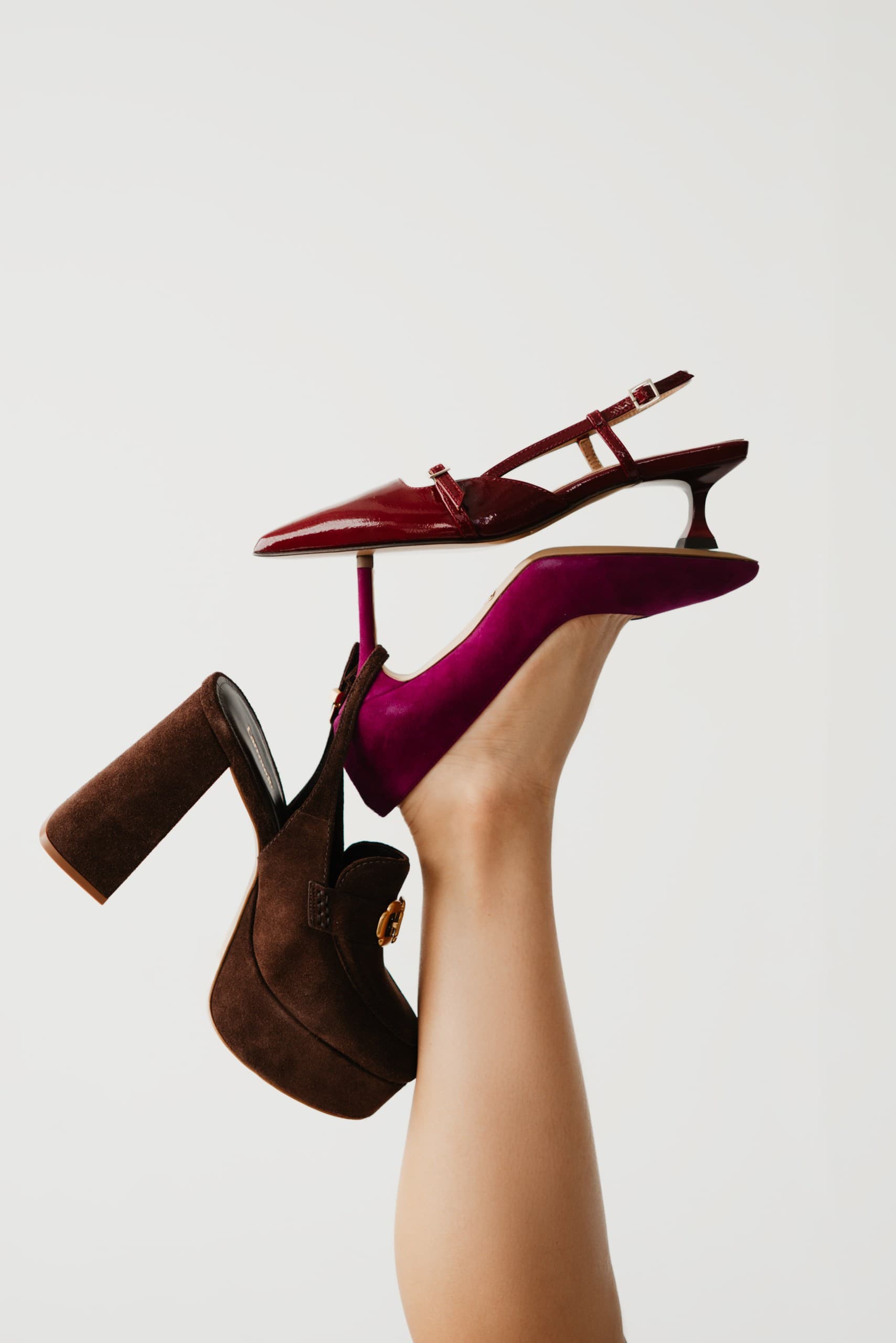 A burgundy patent kitten-heel slingback balanced on a raised leg with a chocolate suede platform pump
