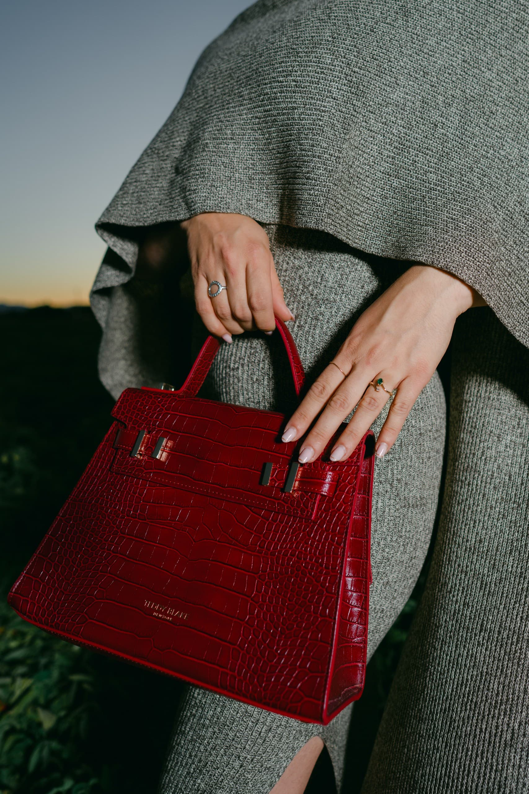A burgundy croc-effect top-handle bag held against a grey knit wrap at sunset