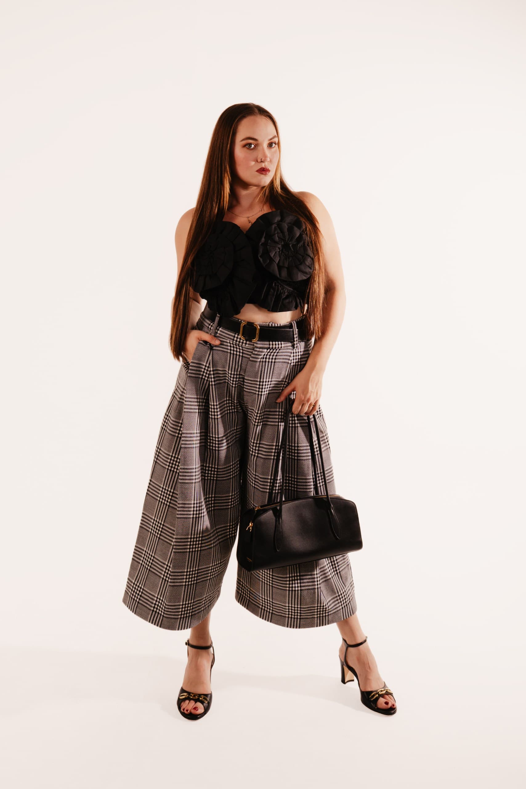 A woman in check wide-leg trousers, a black rosette bustier and black heels, holding a black leather bag