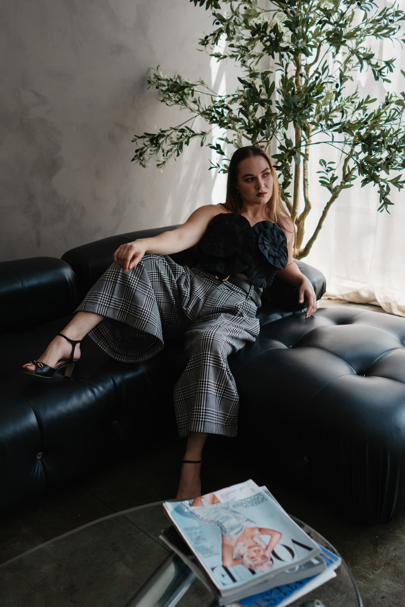 A woman lounging on a black tufted leather sofa in check wide-leg trousers, magazines on a glass table