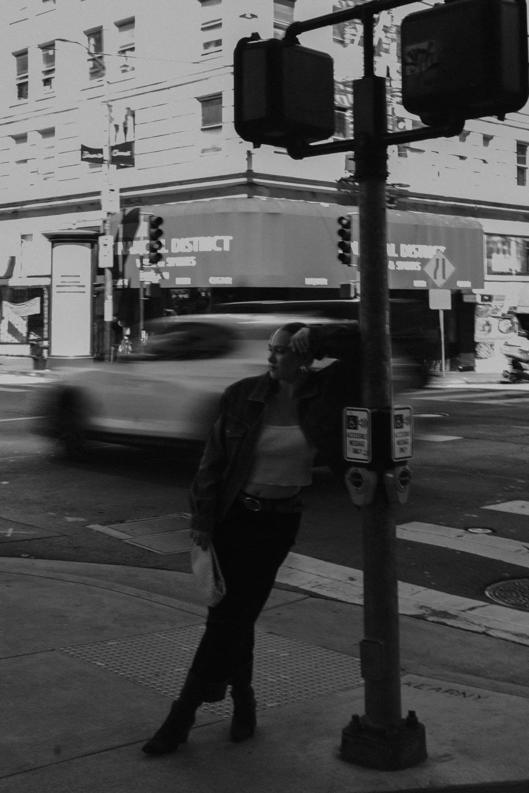 Black-and-white photo of a woman leaning on a traffic-light pole at a city corner as a car blurs past