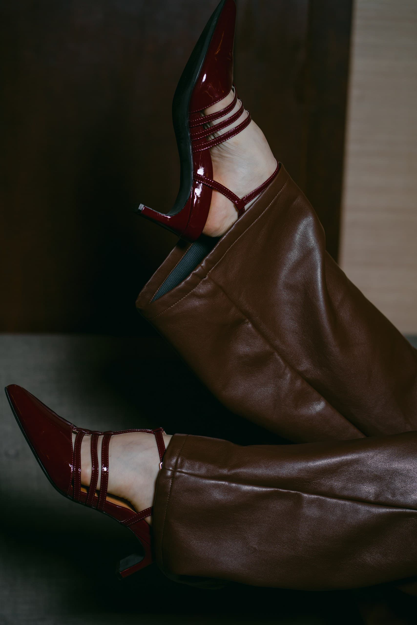Burgundy patent Mary Jane heels and brown leather trousers, legs raised against a dark wall