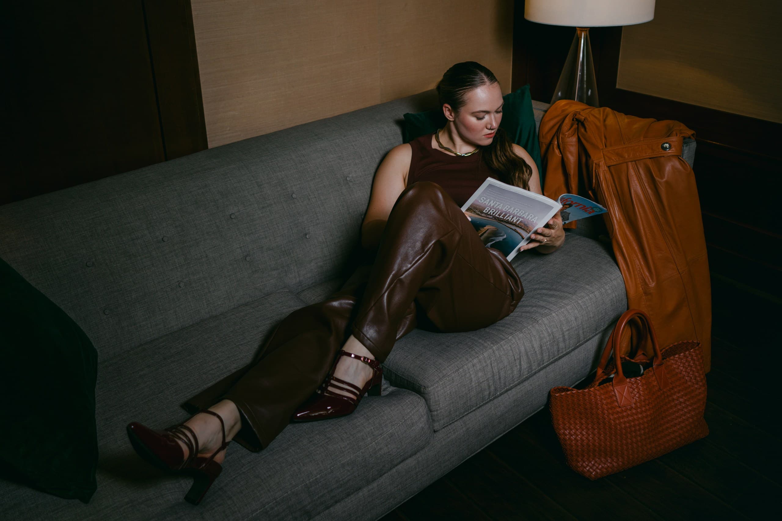 A woman stretched out on a grey sofa reading a magazine in brown leather trousers and red Mary Janes, a cognac coat and woven tote beside her