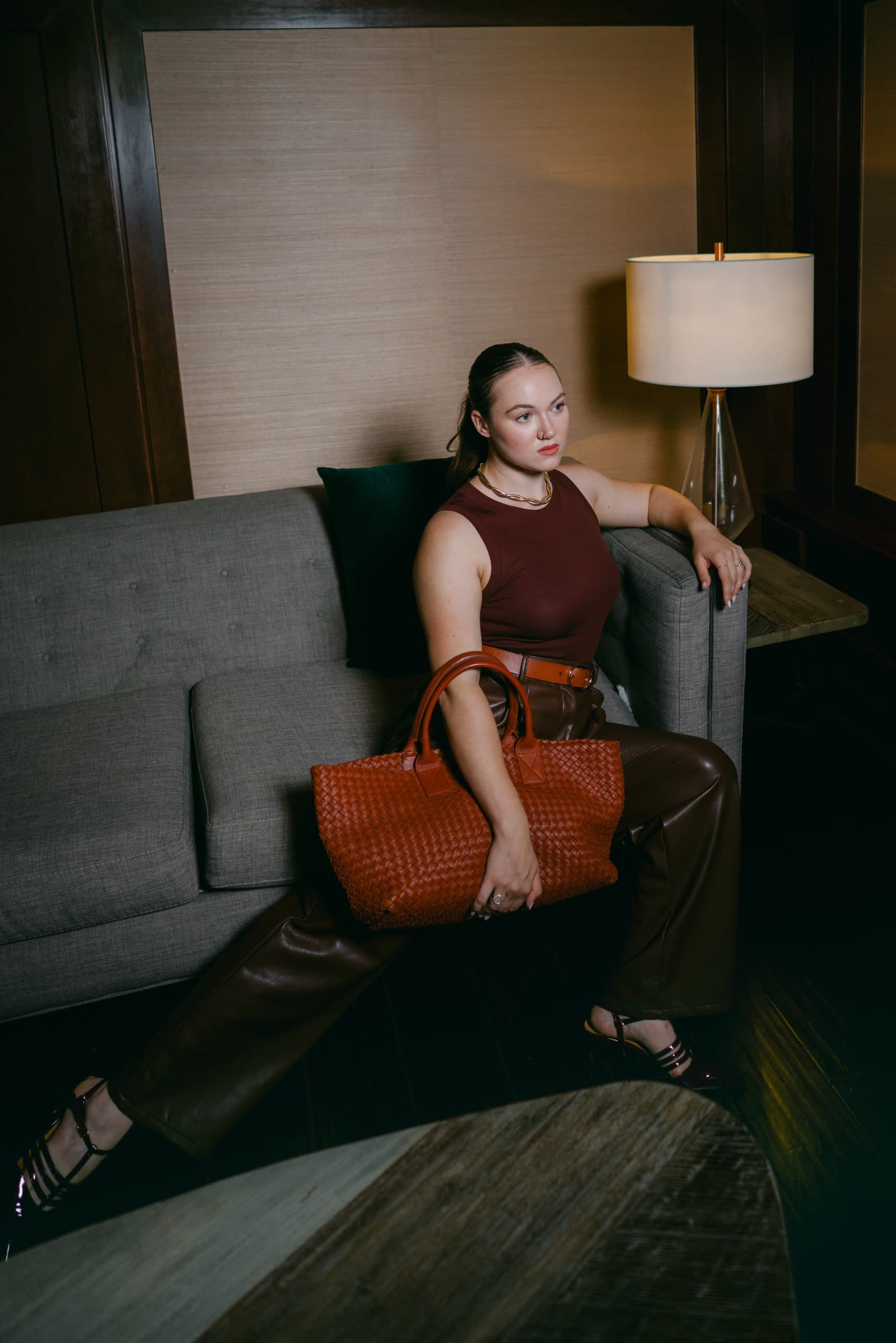 A woman in a burgundy tank and brown leather trousers on a grey sofa, a woven cognac tote on her lap, a lamp glowing behind her