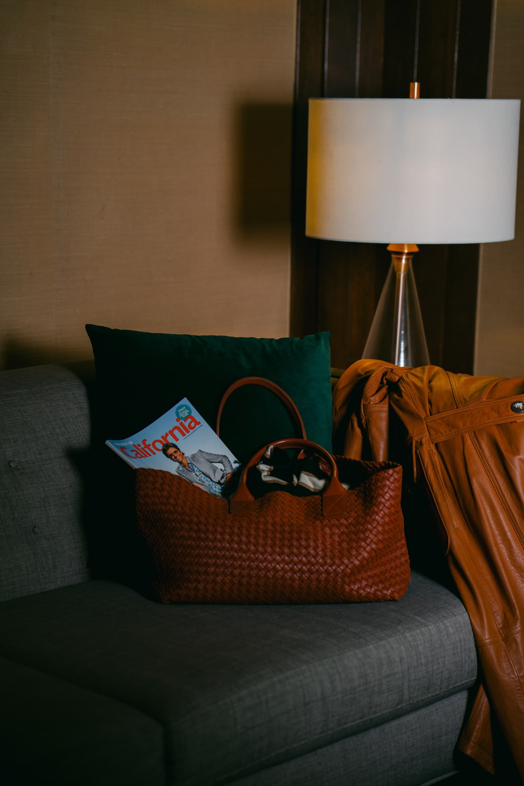 A woven cognac tote with a magazine tucked inside, sitting on a grey sofa under a table lamp